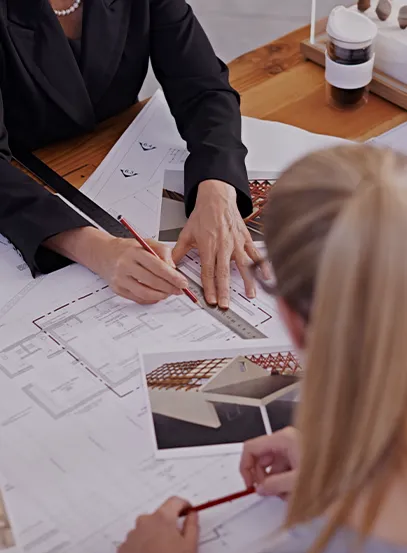 Two professionals sketch architectural plans on a wooden table, using a ruler and pencil amid blueprints and sample photos on the desk.