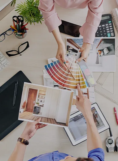 Team of designers examines color swatches and interior photos spread across a bright desk, with a laptop, tablet, plant, and calculator nearby.